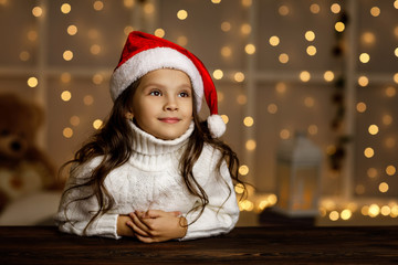Happy smiling little child girl in santa hat dreaming about the holidays on background with lights....