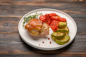 Homemade baked chicken thigh, zucchini and peppers with soy sauce, spices and herbs on a white plate on a wooden background. Close up