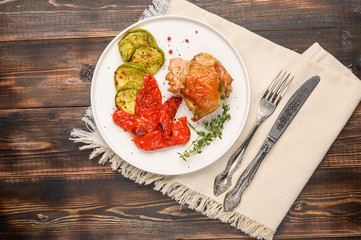 Homemade baked chicken thigh, zucchini and peppers with soy sauce, spices and herbs on a white plate on a wooden background. Top view