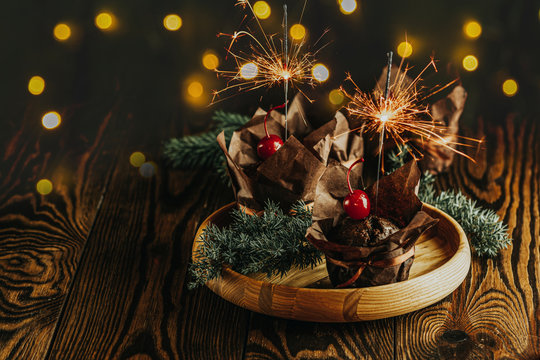 Sweet chocolate muffins with sparklers decorated cherry in brown paper with ribbon on wooden bowl surrounded  pine branches. Close up, shallow depth of the field, greeting food composition.