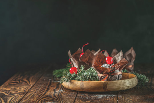 Sweet chocolate muffins decorated cherry in brown paper with ribbon on wooden bowl surrounded  pine branches.