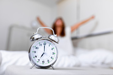 Closeup alarm clock having a good day with background happy woman stretching in bed after waking up, sunlight in morning. Waking up in the morning on time
