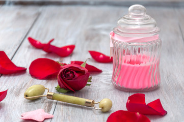 Pink scented candle in a glass jar, Massage, jade roller for the face, a red rose  on a gray wooden table.  Greeting card.  Valentine's Day. Personal care. Relaxation, aromatherapy at home.