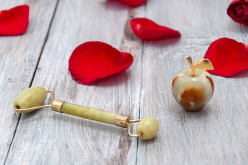 Jade roller for facial massage, jade apple and red rose petals on a gray wooden background. Personal care.