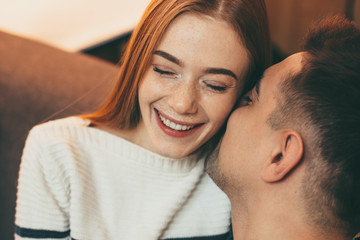 Obraz premium Close up portrait of a lovely female with red hair and freckles laughing with closed eyes while her man is kissing her on the cheek sitting on the couch.