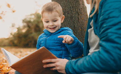 Fototapeta premium Portrait of a cute little boy reading a book outdoor with his mother smiling.