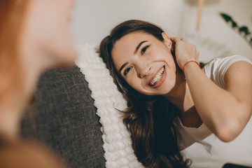 Portrait of a beautiful young woman laughing while leaning head on a bed touching her neck with the head looking to her friend.