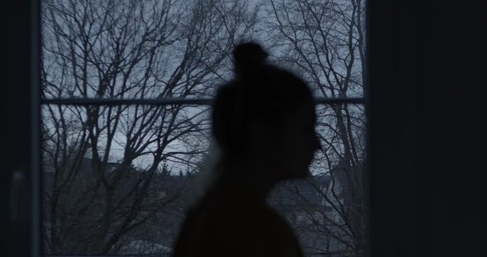 Lonely person in a dreary environment. The silhouette of a young woman looks out the window on a dark day and walks away. Camera from behind. Outside are trees without leaves that sway in the wind.