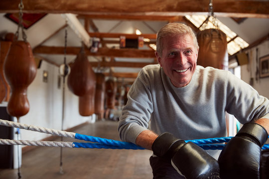 Smiling Senior Male Boxer In Gym Wearing Boxing Gloves Leaning On Ropes Of Boxing Ring