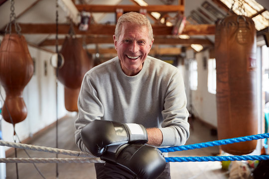 Smiling Senior Male Boxer In Gym Wearing Boxing Gloves Leaning On Ropes Of Boxing Ring