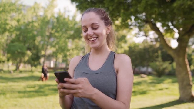 Caucasian fit young woman smiling while texting on mobile phone at the park in the morning on a sunny day