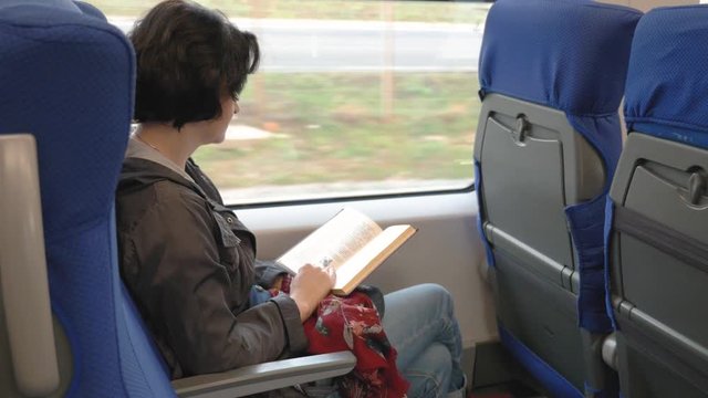 at the window of a moving train sits Caucasian a middle aged woman with short hair brunette in a black jacket blue jeans Reading a book side View