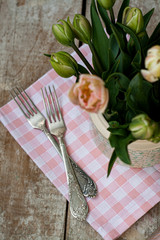 Two forks lie on a pink napkin next to a vase of delicate pink tulips. The composition stands on a wooden old background.