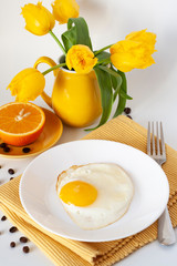 Bright breakfast: egg, juicy orange, bouquet of yellow tulips, yellow napkin and fork. The whole composition stands on a white background.