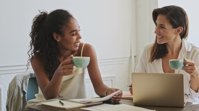 Happy young beauty women colleagues working with documents and laptop computer while drinking coffee and sitting by the table at office