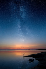 a man standing by a calm water and looking at the stars of milky way on the sky with reflections...