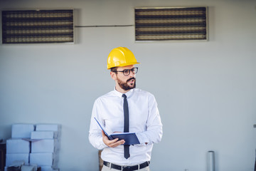 Strict caucasian director in shirt and tie, with eyeglasses and helmet on head standing in front of printing shop and controlling workers.