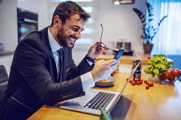 Fototapeta na wymiar Smiling attractive caucasian businessman in suit sitting at dining table and using smart phone for reading or sending message. On table are laptop, dish and vegetables.