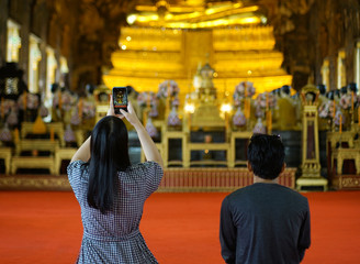 Young Asian couples tourists wear casual clothes worship and taking photo of golden Buddha statue inside chapel of Buddhist temple in Thailand by smartphone camera.