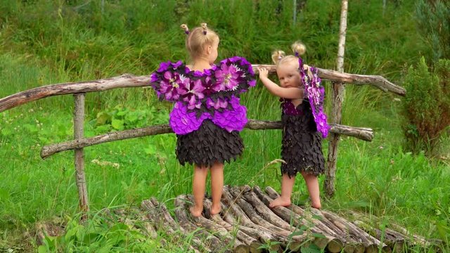 Two blond girls with butterfly wings made of flowers are standing on a wooden bridge. Children pretend to be purple butterflies