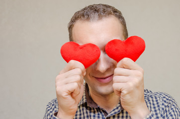 Obraz premium attractive dark-haired man with a smile, with two small red hearts in his hands, covering his eyes with paper heart symbols, standing on a light background. Valentine's day