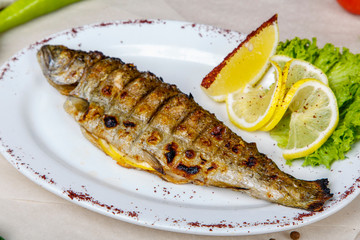 grilled trout with lemon and lettuce on a white plate. The plate is on white and gray wrapping paper, lined with pepper, peas, tomatoes, garlic, herbs