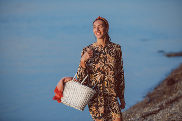 Slim attractive caucasian brunette in floral dress holding picnic basket and walking on the shore.