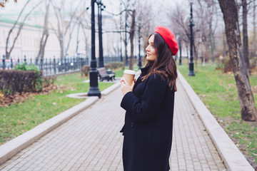 Beautiful young girl in a black coat and red beret hold hot drink in a craft plastic cup with a...