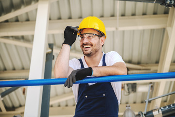 Smiling handsome caucasian worker in overall and with helmet on head leaning on the railing. Oil...