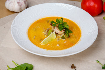 Cheese soup with shrimp. Yellow soup is decorated with lemon and parsley. White round plate lies on a white and gray wrapping paper, overlaid with pepper, peas, tomatoes, garlic, herbs. 