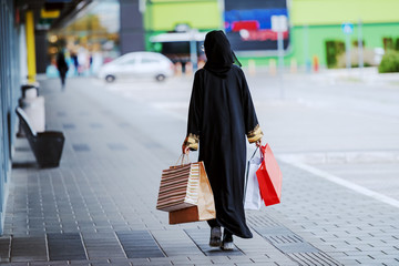 Rear view of Muslim woman in traditional wear walking outdoors with shopping bags in hands. Fashion is for everyone. Diversity concept.