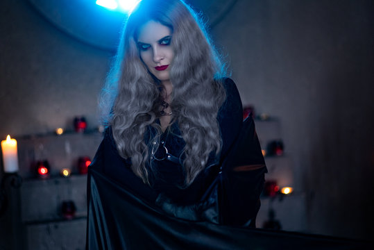 Spooky woman in witch costume posing in dark gothic building with candles on background
