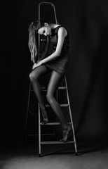 Beautiful brunette woman in black dress posing while sitting on ladder in dark studio