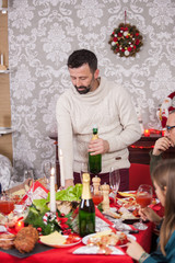 Cheerful adult man pops a bottle of champagne celebrating christmas with his family