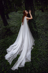 Fototapeta premium Attractive ginger elf woman in white dress and cape leaning on tree while posing in green forest