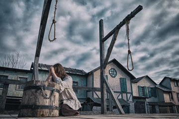 A woman in medieval dress prays against a cloudy dramatic sky. Place of execution with gallows and scaffold on the background of old houses - obrazy, fototapety, plakaty