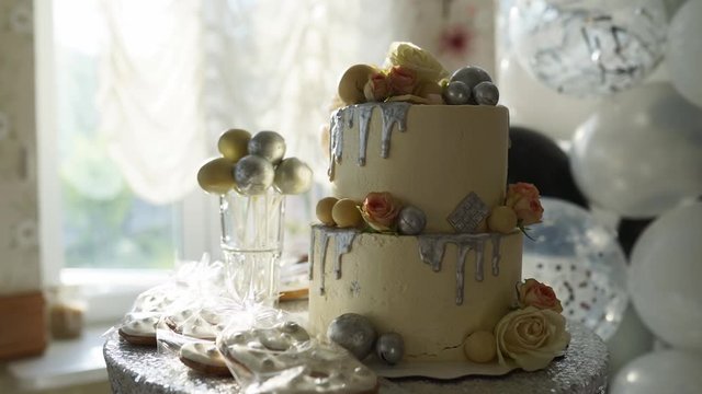 Wedding cake with roses, white glaze and silver topping on candy bar. Cakepops, biscuits and cookies on birthday party served table. Balloons on background.
