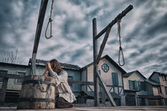 A woman in medieval dress prays against a cloudy dramatic sky. Place of execution with gallows and scaffold on the background of old houses
