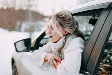 A beautiful attractive blonde in a white warm sweater travels and looks out of the car window at the snow covered road against the background of a snowy winter forest