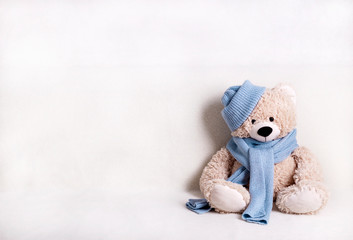 Children's toy teddy bear is sitting on a white sofa in a knitted scarf and hat in blue.
