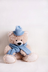 Children's toy teddy bear is sitting on a white sofa in a knitted scarf and hat in blue.