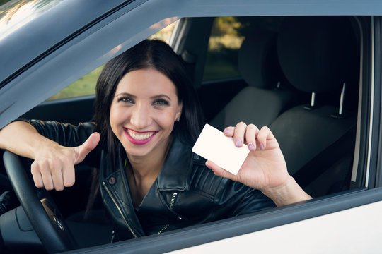 Young girl with beautiful smile sitting in white car showing an empty white card and thumbs up hand sign. Woman has got driving license and is very happy. Pretty girl at wheel inside car is excited.