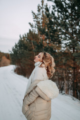 Happy attractive beautiful lonely blonde in a white sweater, warm jacket and jeans walks, enjoys freedom and tranquility on a snowy road in the forest in winter.