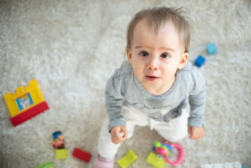 Portrait of one year old baby sitting on carpet in bright room looking up towards camera. Copy space on left.