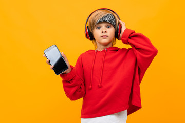 European blond boy in a red hoodie listens to music in red headphones and holds smartphone with mockup on a orange background