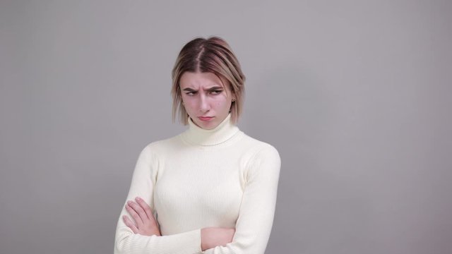 Portrait of angry young woman looking aside and covering chin with hand isolated on gray wall background in studio. People sincere emotions, lifestyle concept.