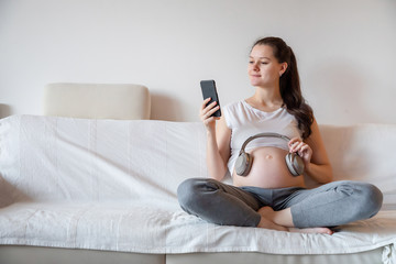 Pregnant woman sitting on white sofa at home and keeping headphones on her belly