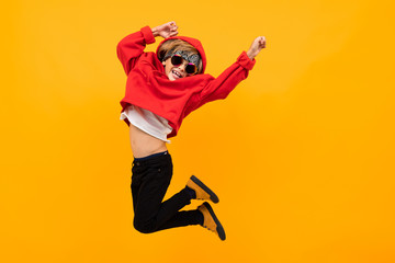 handsome boy with a bandana on his head in a red hoodie with glasses jumps on an isolated orange...