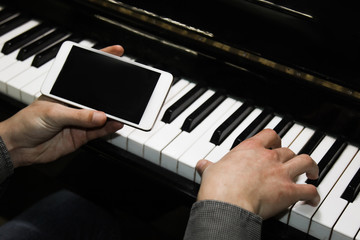 Fototapeta na wymiar two male hands on the piano with a smartphone. palms lie on the keys and play the keyboard instrument in a music school. student learns to play. hands of a pianist. black dark background.