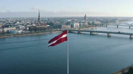 Latvian flag attached to a pole waving in the wind in front of the Daugava river and the skyline of Riga, Latvia